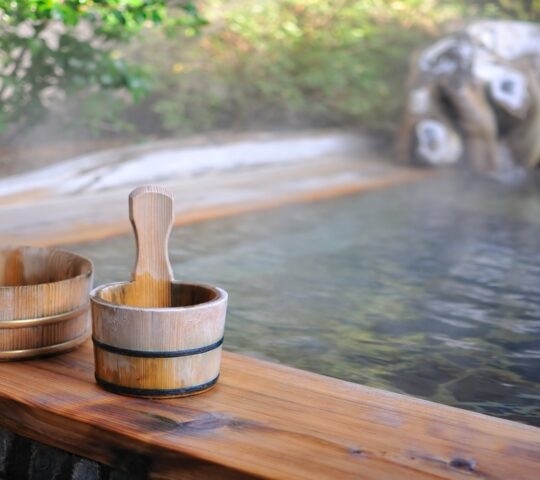 Wooden water buckets rest on the edge of a steaming outdoor onsen bath with a stone and foliage background.