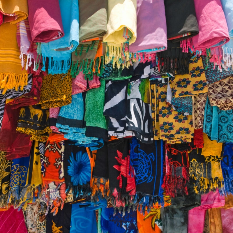 Luxury Kenya Safari Tours - Rows of brightly colored, patterned fabrics with fringes hanging vertically in a dense marketplace arrangement.