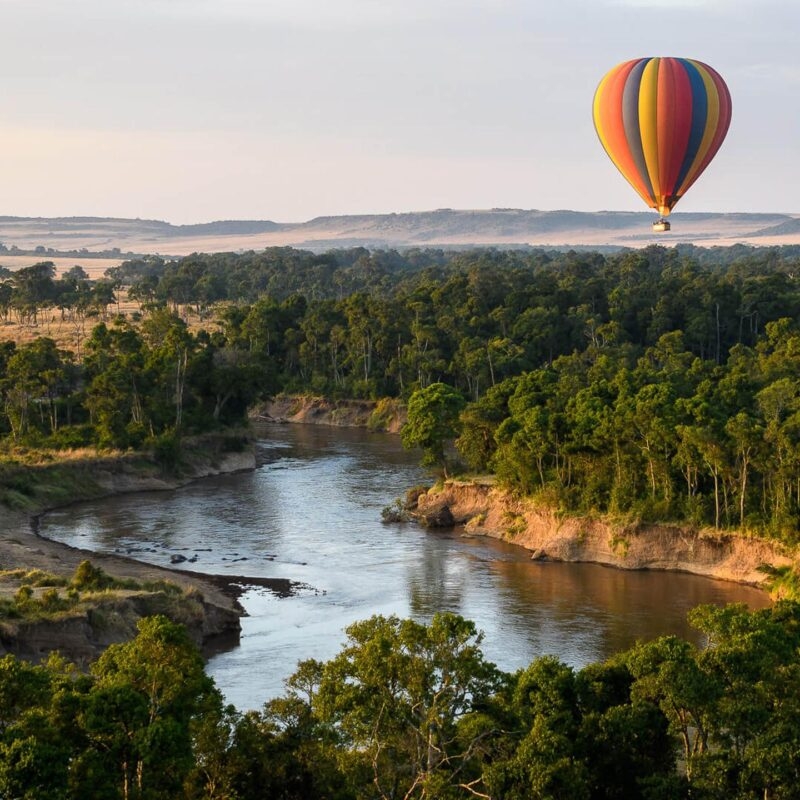 Luxury Kenya Safari Tours - A colorful hot air balloon flying over a river lined with green trees under a clear sky.