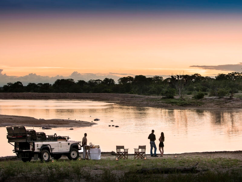 Luxury South African Tours - People stand by a calm lake at sunset next to a white safari vehicle and a table with white cloth.