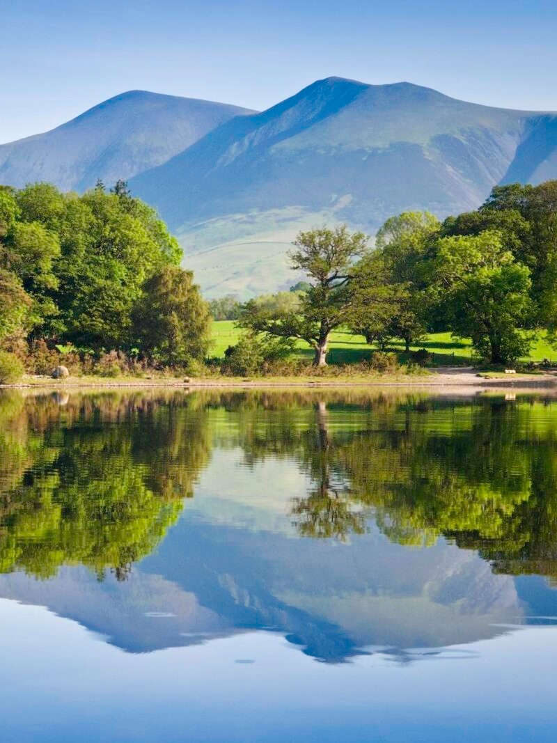 Calm lake reflecting green trees and a large, smooth mountain under a bright blue sky in a pastoral landscape.
