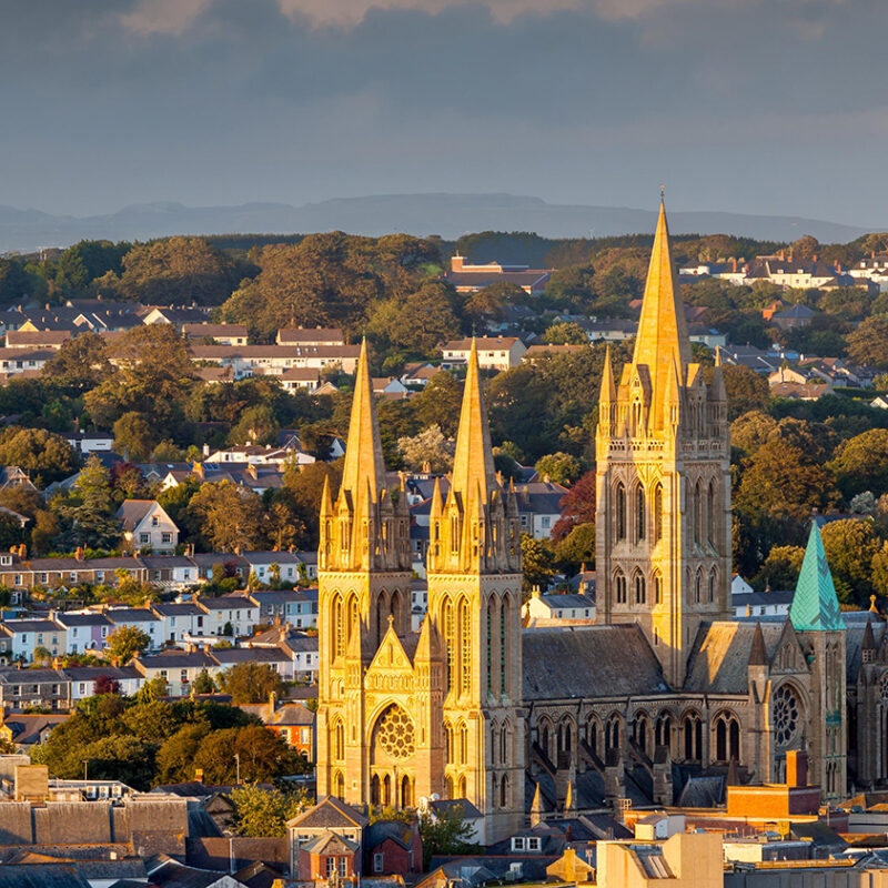 The sunlit, three-spired Truro Cathedral dominating the view over the surrounding houses and green trees of Cornwall.