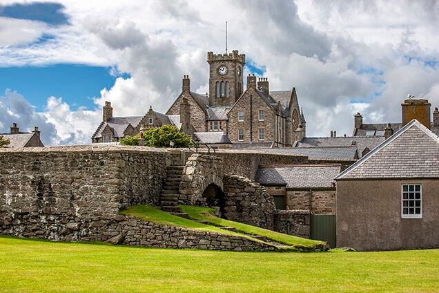 Stone building with a clock tower and a traditional stone boundary wall with stairs leading to an archway.