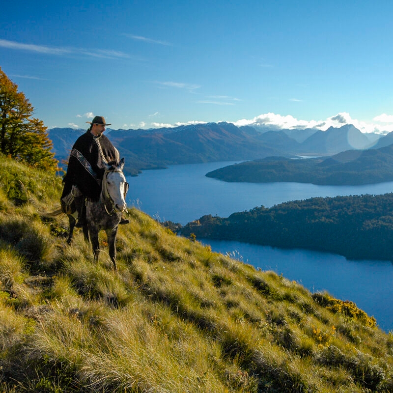 A couple enjoying some spectacular views near Bariloche in PatagA couple enjoying some spectacular views near Bariloche in Patag - A person riding a horse on a high grassy path above a blue lake on a luxury Patagonia tour