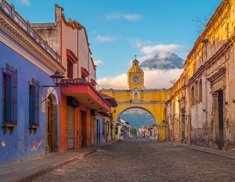The yellow Santa Catalina Arch stands over a cobblestone street in Antigua, Guatemala, with a volcano visible in the distance.
