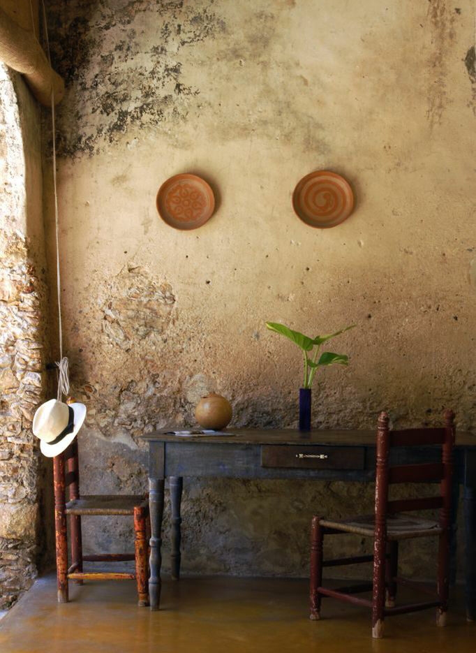 Desk and chairs against a textured stone wall with a hanging straw hat and two plates, representing luxury Mexico trips.