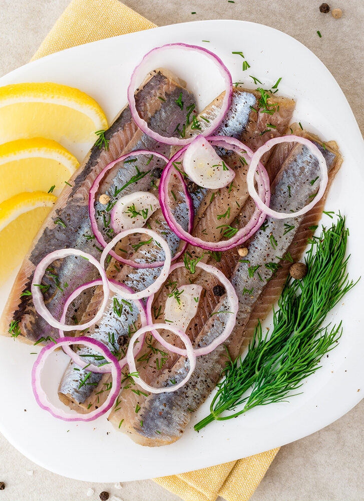 Pickled herring fillets on a white plate, garnished with red onion rings, dill, and lemon wedges, typical for luxury Norway trips.