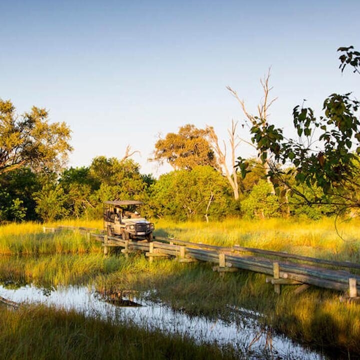 A white safari vehicle driving across a rustic wooden bridge over a small stream in a grassy field.