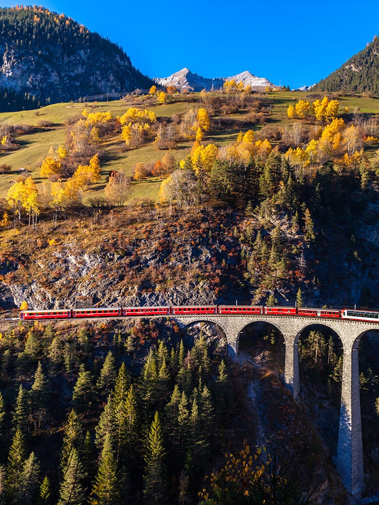 Iconic red train crossing a high arched stone bridge surrounded by golden autumn trees on luxury Switzerland trips.