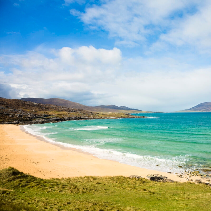 A wide, sandy beach with turquoise water and waves breaking onto the shore, surrounded by grassy hills and blue sky. Ideal for luxury Scotland trips.
