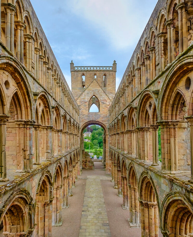 A long corridor view between two rows of symmetrical stone arches inside the ruins of Jedburgh Abbey. A great stop on luxury Scotland trips.