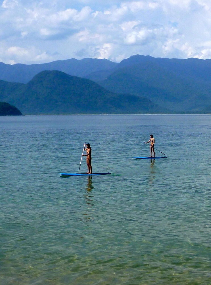 Two women stand up paddleboarding in a bay with mountains in the background. luxury Brazil trips.