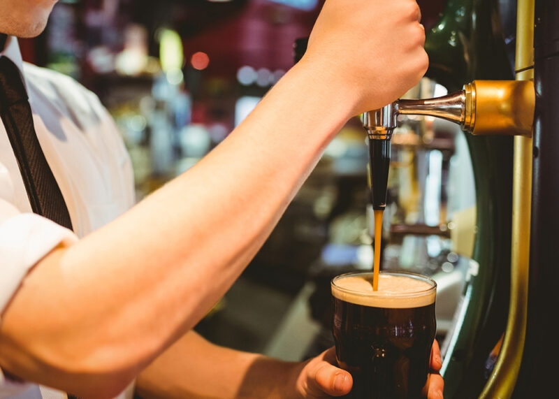 Close-up of a bartender's hands pouring a dark stout beer from a brass tap into a pint glass in a pub. Experience local flavors on luxury Ireland tours.