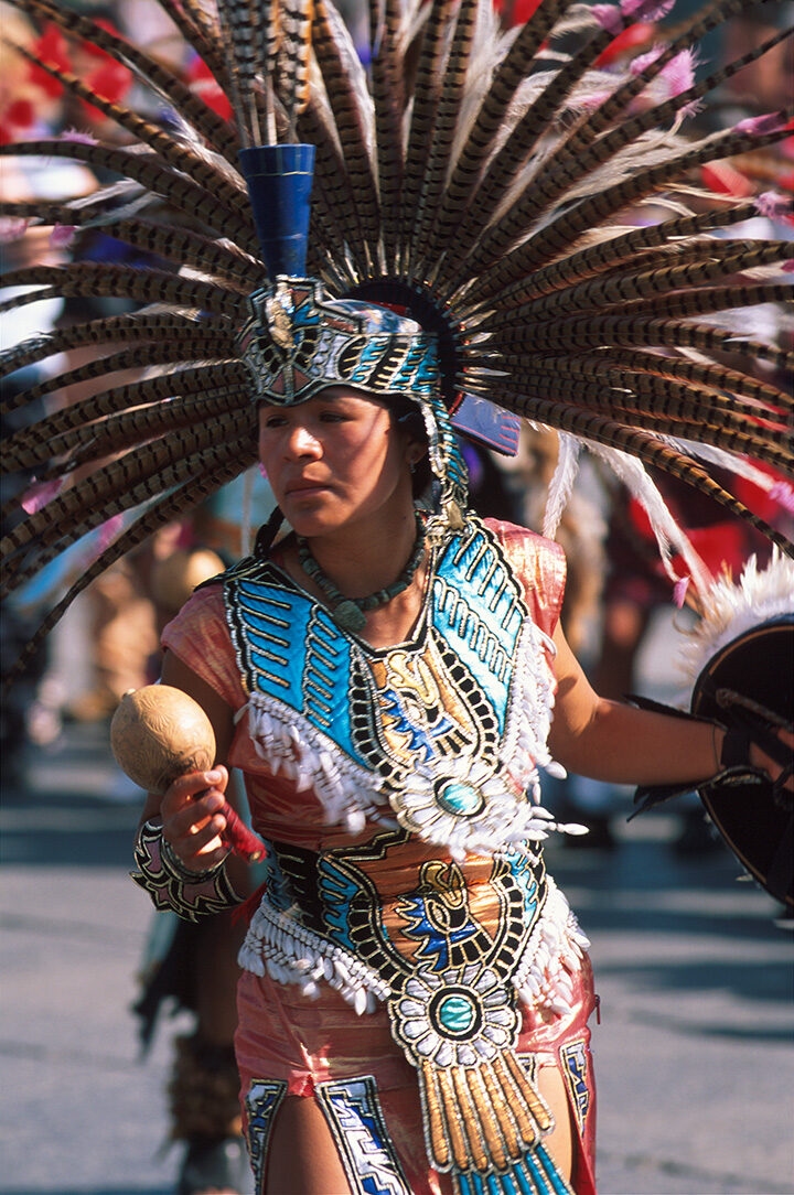 A woman in full traditional Aztec-style feathered headdress and ceremonial attire holding a maraca, luxury Latin America grand tour holidays.