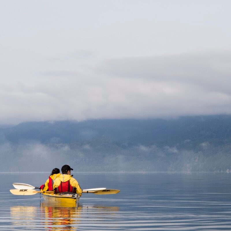 Two people in a yellow kayak paddling on still water in front of misty mountains