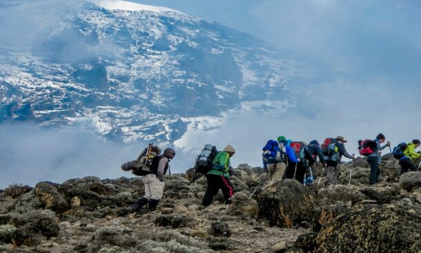 A line of hikers with gear climbs a rocky slope toward a large, snow-capped mountain.