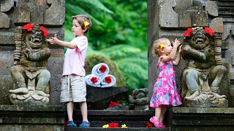 A young boy and girl standing by ornate stone statues decorated with red flowers in a tropical garden setting.