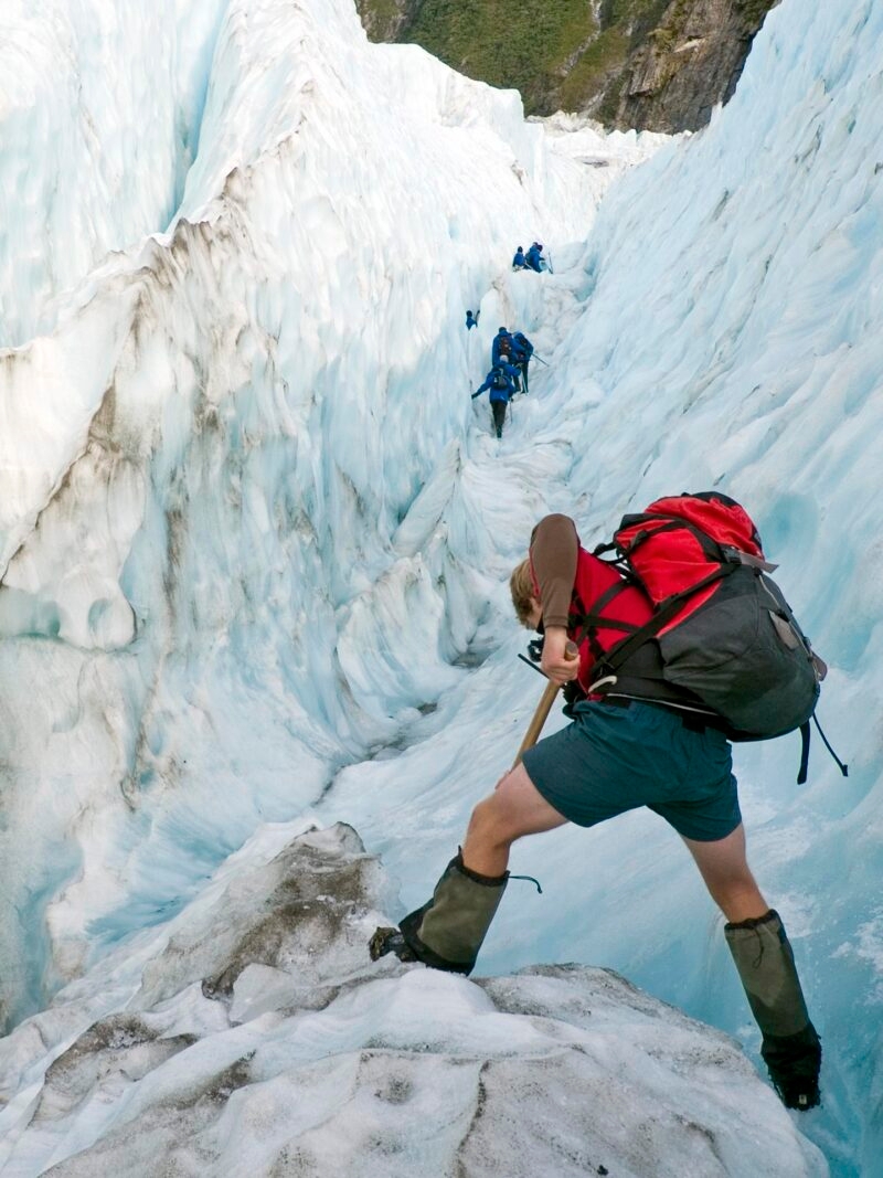A hiker with a red backpack walks down a rocky mountain path on epic expeditions in New Zealand.