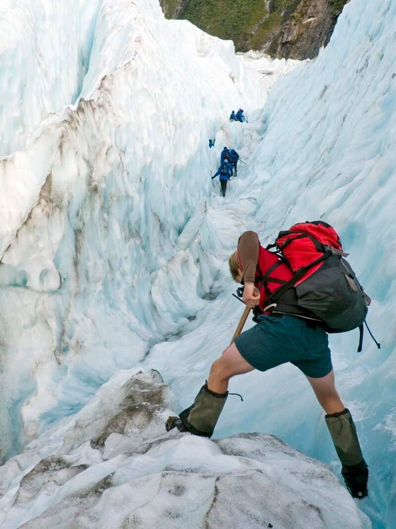 A hiker with a red backpack walks down a rocky mountain path on epic expeditions in New Zealand.