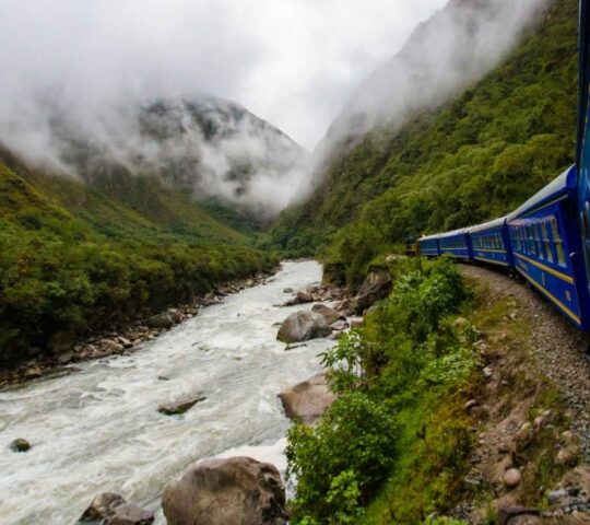 Blue passenger train rounding a curve next to a rocky, rapid river in a fog-covered, verdant mountain valley.