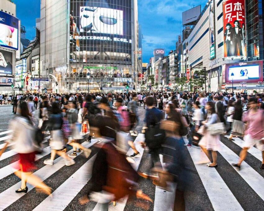 A wide, blurry shot of massive crowds of people crossing the street at Tokyo's Shibuya intersection at dusk.