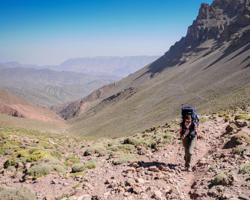 Female Hiker walking up a mountain pass with backpack on her back on a single footpath up a mountain with a valley and a clear blue sky behind Atlas Mountain Toubkal Morocco