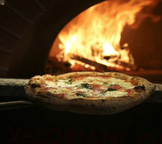 Margherita pizza on a peel being removed from a wood-burning oven with bright orange flames inside.