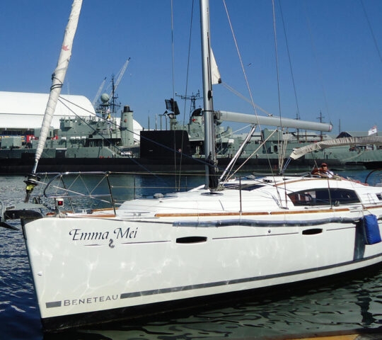 A white Beneteau sailing yacht named Emma Mei docked in a harbor with naval vessels in the background.