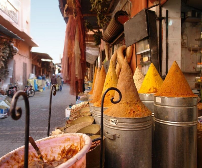 Conical piles of orange and yellow spices stand in silver containers at an outdoor market.