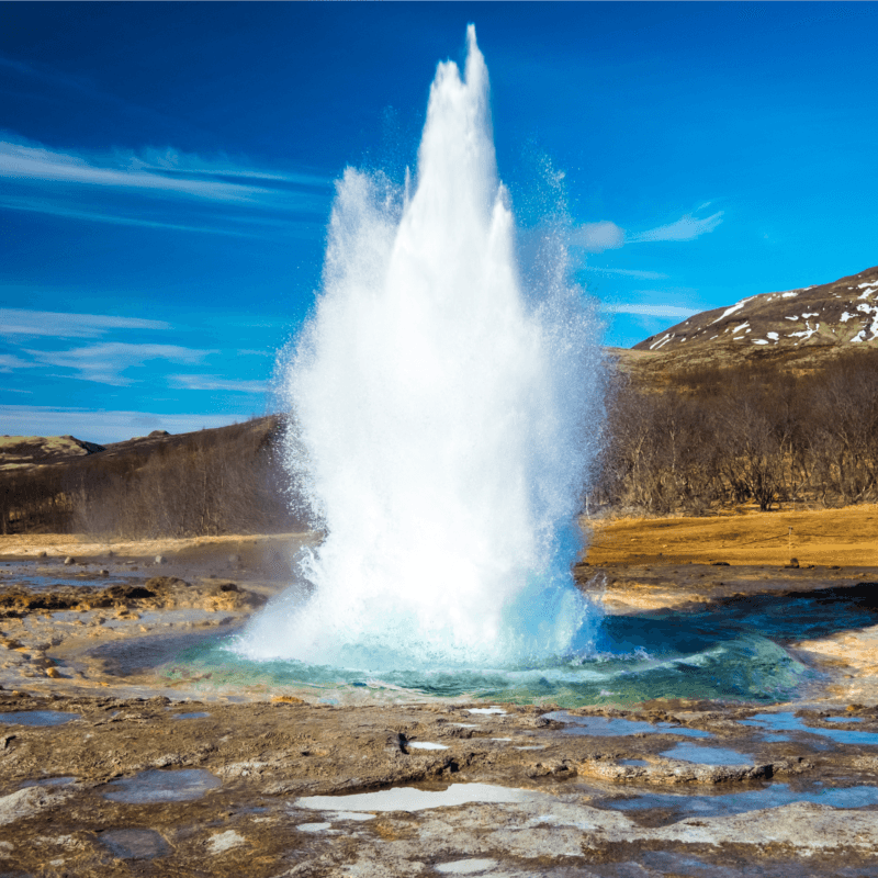 A white geyser erupts from a pool of blue water during luxury golden circle tours, set against hills and a blue sky.