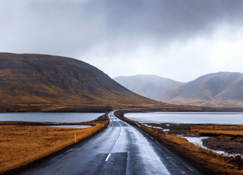 A long, winding road beside a lake and mountains under a cloudy sky during luxury Golden Circle tours.