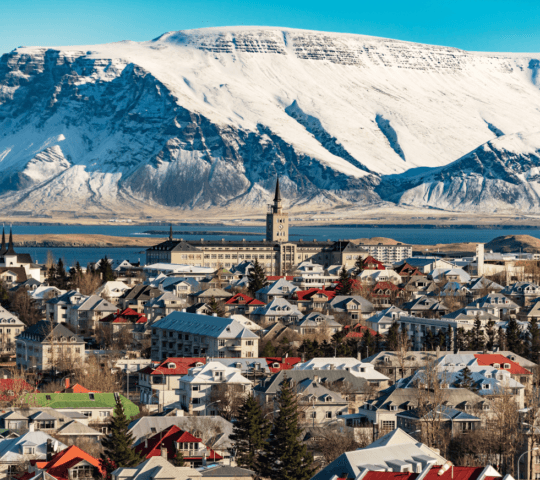 A snowy mountain backdrop behind a town with distinct architecture and a tall spire.