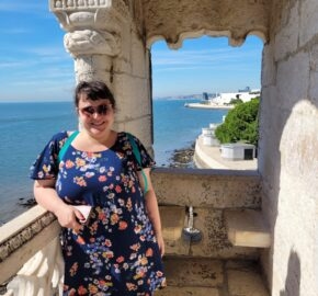 Person in floral dress standing in stone archway overlooking sea and coastline.