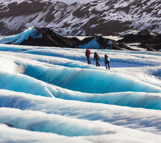 Three hikers traverse a vibrant blue glacier with dark mountains in the background.