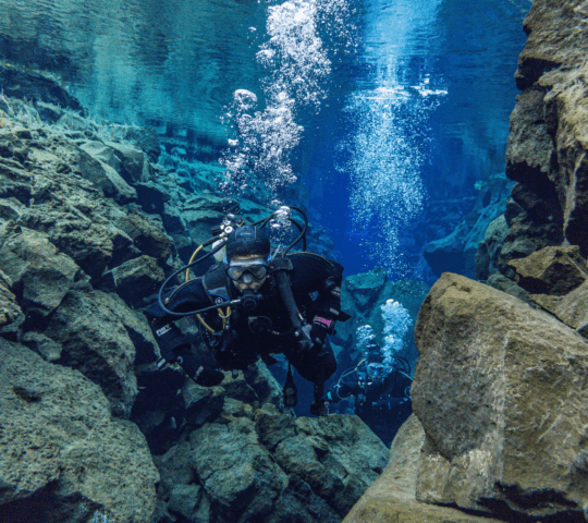 A person scuba diving in a wetsuit in clear water with rocks around