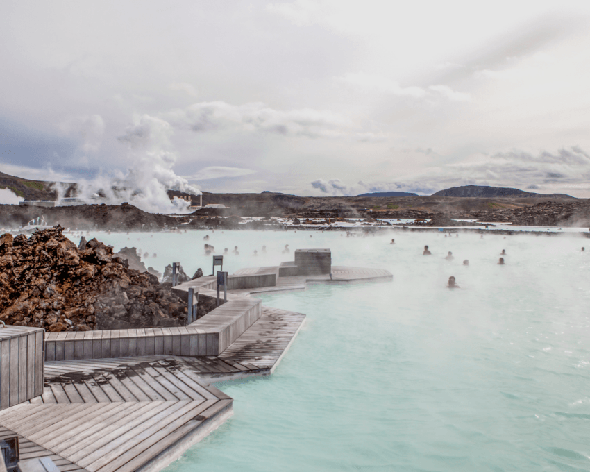 Visitors enjoy a geothermal spa with steamy blue waters amidst rocky terrain under a cloudy sky.