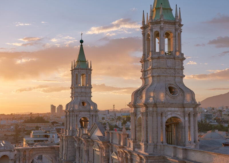 Twin bell towers of a cathedral during sunset with a cityscape backdrop.