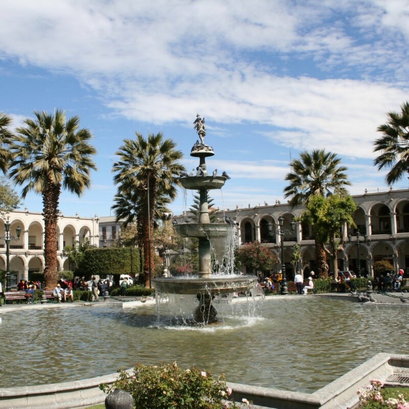 A sunny day at a plaza with a central fountain, palm trees, and surrounding colonnaded buildings.