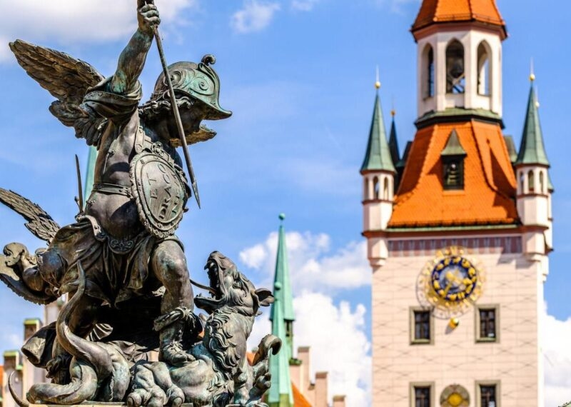 Bronze statue of a winged figure in front of a clock tower with a blue sky background.