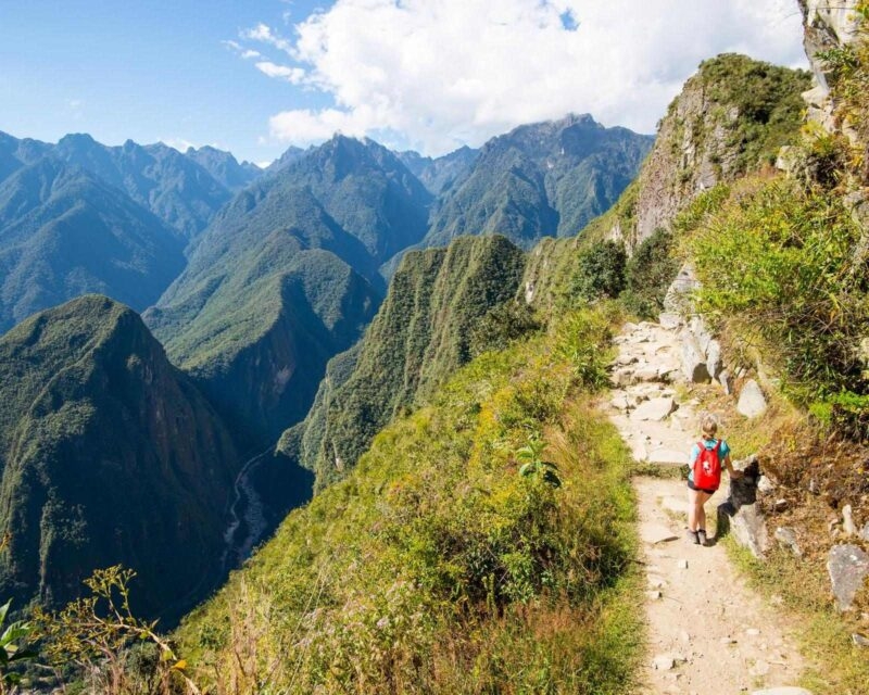 A person in a red backpack hikes on a narrow mountain path overlooking a deep, lush green valley and towering mountains, a highlight of luxury Machu Picchu holidays.