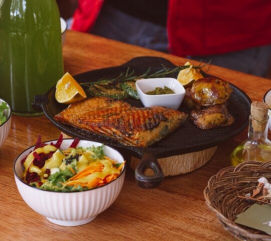 A spread of grilled fish, salads, roasted potatoes, soup, and a green bottle on a wooden table.