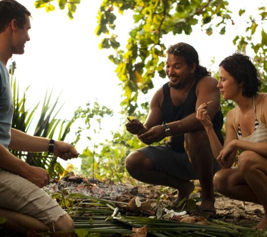 A group of three people crouched together outdoors, looking at items spread on large green leaves on the ground.