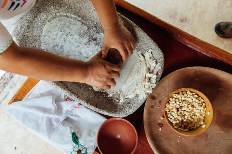 Close-up of hands grinding corn dough on a large, grey stone metate, with a bowl of dried corn kernels nearby. For luxury Belize tours.