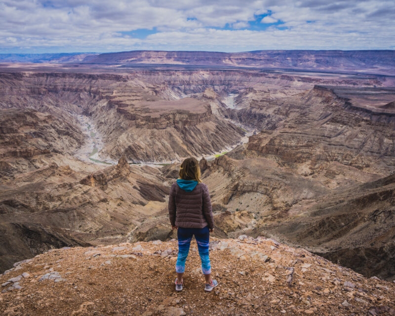 Hiker overlooking the deep, rocky ravines and winding paths of Fish River Canyon under a cloudy sky.