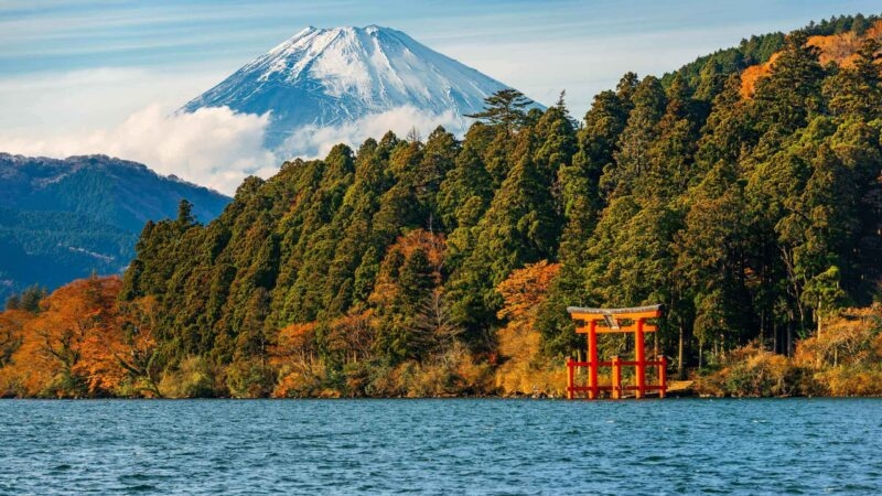 Red torii gate on the water's edge of Lake Ashi backed by green trees and snow-capped Mount Fuji under a clear sky.