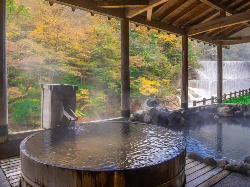 Steaming Japanese hot spring wooden bath overlooking autumnal trees and a waterfall.