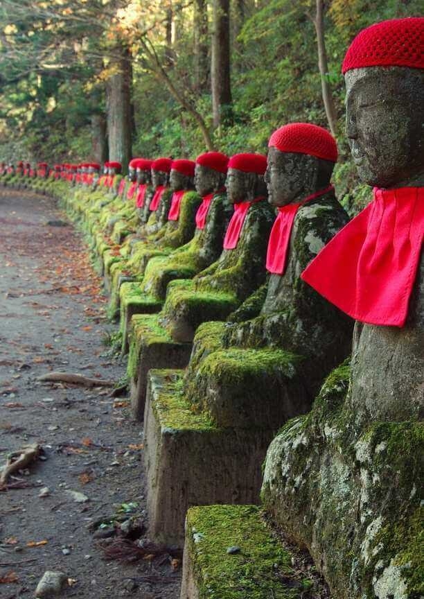 A long row of mossy stone Jizo statues wearing red caps and bibs alongside a forest path.