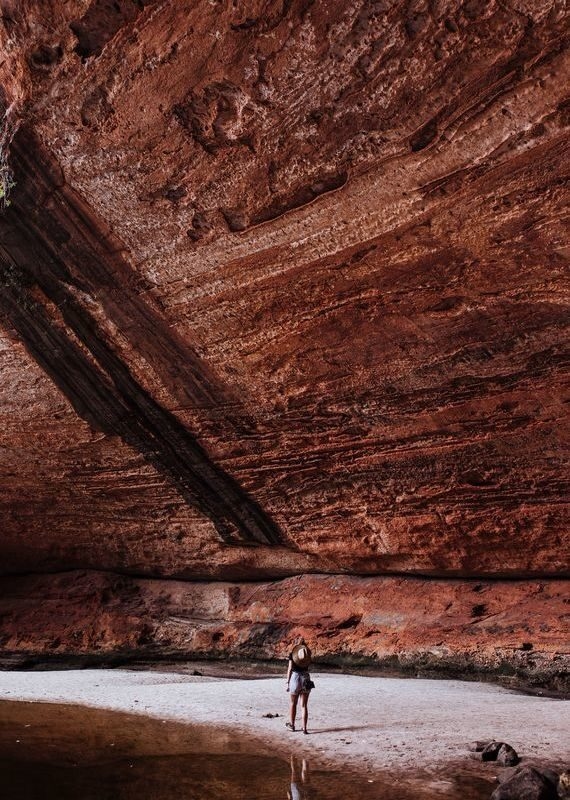 A person stands on a sandy shore beneath a massive layered red rock cave overhang in Australia.