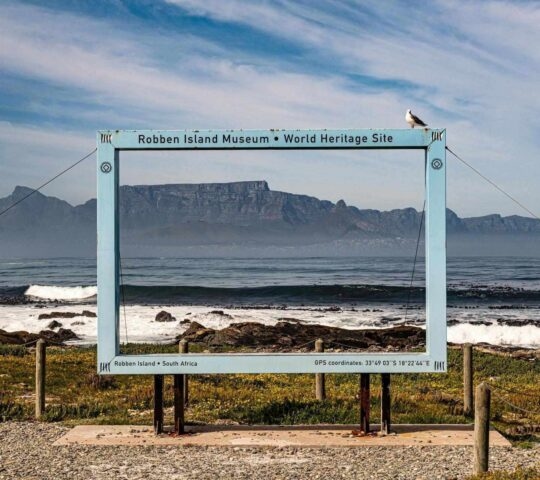 A blue picture frame outdoors on Robben Island overlooks the ocean, framing Table Mountain under a partly cloudy sky.