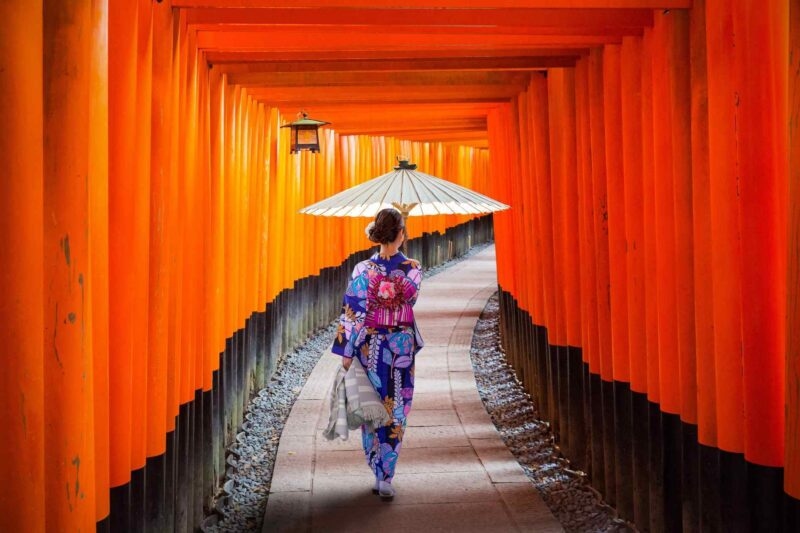A woman in a blue kimono and umbrella walks away along a stone path framed by red torii gates.